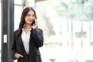 feliz joven empresaria asiática con traje sosteniendo un teléfono móvil de pie en la oficina de su estación de trabajo foto