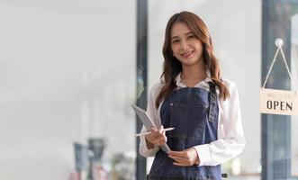 hermosa joven barista asiática en delantal sosteniendo una tableta y parada frente a la puerta de la cafetería con un letrero abierto. concepto de inicio de propietario de negocio.