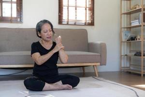 60s aged Asian woman massaging her wrist, feeling pain and swelling in the joints, injured hand during yoga practice at home. photo