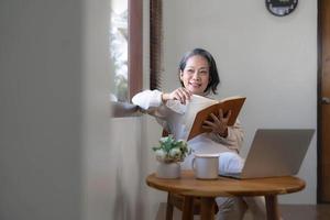 Relaxed and calm 60s retired Asian woman looking out the window, daydreaming while reading a book in her home living room. lifestyle concept photo