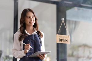 hermosa joven barista asiática en delantal sosteniendo una tableta y parada frente a la puerta de la cafetería con un letrero abierto. concepto de inicio de propietario de negocio.