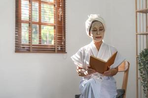 bella dama asiática madura en albornoz con mascarilla de arcilla hidratante facial o antienvejecimiento en la cara está leyendo un libro en la sala de estar. foto