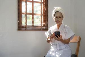 bella dama asiática madura en albornoz con mascarilla de arcilla hidratante facial o antienvejecimiento en la cara está usando su teléfono móvil en la sala de estar.