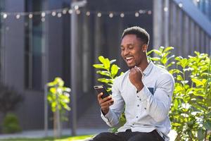 hombre afroamericano feliz con ropa informal sentado en un banco al aire libre con teléfono móvil