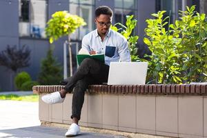 Young african american male student sitting in a city park on a bench with laptop and notebook photo