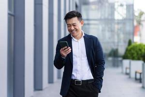 autónomo masculino asiático caminando cerca del centro de negocios sosteniendo el teléfono, sonriendo leyendo noticias foto