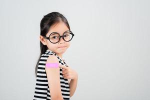 Asian little girl wear glasses showing her arm with bandage after virus vaccine happy Kid and child vaccination campaign. Protect your kid with vaccine concept photo