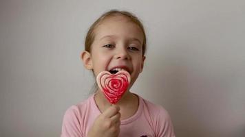 A cute little girl licks a big red heart-shaped lollipop. Isolated white background. video
