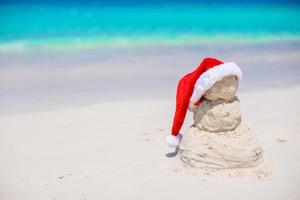 pequeño muñeco de nieve de arena con sombrero rojo de santa en la playa caribeña blanca foto