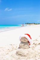 muñeco de nieve de arena con sombrero rojo de santa en la playa caribeña blanca foto