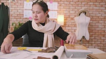 asiatisk medelålders kvinna mode designer Arbetar i studio, skärande och välja tyg mönster idéer med teckning skisser för klänning design samlingar. professionell boutique skräddare sme entreprenör. video
