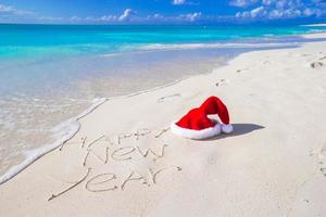 feliz año nuevo escrito en la playa de arena blanca con sombrero rojo de santa foto