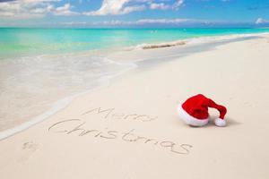 sombrero de santa en la playa de arena y feliz navidad escrito en arena blanca foto