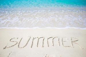 palabra verano escrita a mano en una playa de arena con una suave ola oceánica en el fondo foto
