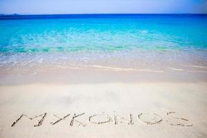 Word Mykonos handwritten on sandy beach with soft ocean wave on background photo