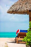 Santa Claus Hat on chair near tropical beach with turquoise sea water and white sand. Christmas vacation concept photo