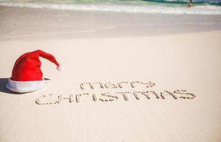 sombrero de santa en la playa de arena blanca y feliz navidad escrito en la arena foto