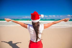 vista trasera de una hermosa chica con sombrero de santa caminando extendió sus manos en la playa blanca foto