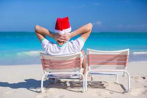 vista trasera del joven con sombrero de navidad en la silla de playa foto