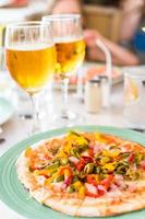 Pizza with mozzarella cheese, olive, fresh tomato and pesto sauce. Served at restaurant table with two beers photo