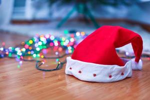 Red Santa Claus hat on a timber floor with brightly colored garlands, in lights background.