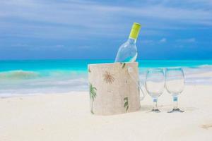 botella de vino blanco y dos copas en la exótica playa de arena foto