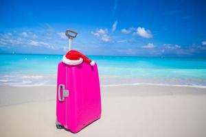 Pink suitcase and Santa Claus hat on white sand beach photo