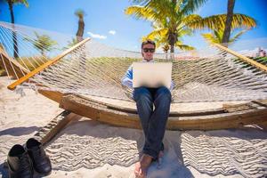 Businessman sitting in hammock using laptop during summer vacation photo