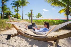 Young man working on laptop in hammock at tropical beach photo
