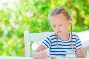 Adorable little girl having breakfast at cafe with sea view early in the morning photo