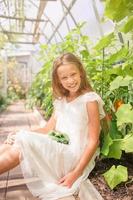 adorable niña cosechando pepinos y tomates en invernadero. retrato de niño con cesta con verduras foto