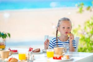 Adorable little girl having breakfast at cafe with sea view early in the morning photo