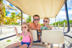 papá y sus niñas conducen un carrito de golf en una playa tropical foto