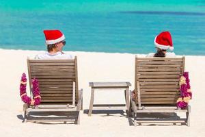 Vista trasera de una pareja joven con sombreros de Papá Noel relajándose en una playa tropical durante las vacaciones de Navidad foto