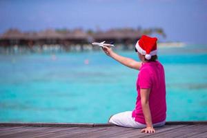 joven con sombrero de santa durante las vacaciones en la playa en un embarcadero de madera con una miniatura de avión foto