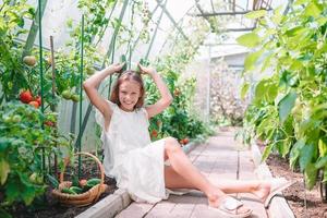 adorable niña cosechando pepinos y tomates en invernadero. foto