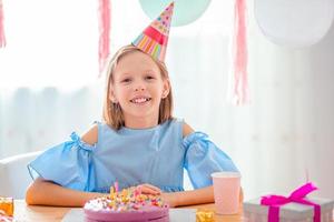 Caucasian girl is dreamily smiling and looking at birthday rainbow cake. Festive colorful background with balloons. Birthday party and wishes concept. photo