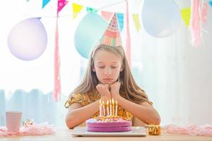 Caucasian girl is dreamily smiling and looking at birthday rainbow cake. Festive colorful background with balloons. Birthday party and wishes concept. photo