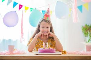 la chica caucásica sonríe soñadoramente y mira el pastel de arco iris de cumpleaños. fondo colorido festivo con globos. concepto de fiesta y deseos de cumpleaños. foto