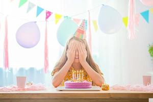 Caucasian girl is dreamily smiling and looking at birthday rainbow cake. Festive colorful background with balloons. Birthday party and wishes concept. photo