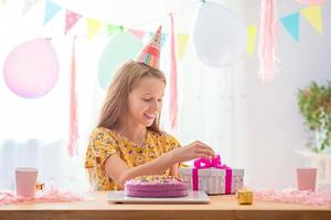 Caucasian girl is dreamily smiling and looking at birthday rainbow cake. Festive colorful background with balloons. Birthday party and wishes concept. photo