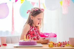 Caucasian girl is dreamily smiling and looking at birthday rainbow cake. Festive colorful background with balloons. Birthday party and wishes concept. photo