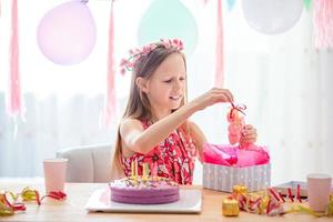 Caucasian girl is dreamily smiling and looking at birthday rainbow cake. Festive colorful background with balloons. Birthday party and wishes concept. photo