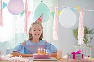 Caucasian girl is dreamily smiling and looking at birthday rainbow cake. Festive colorful background with balloons. Birthday party and wishes concept. photo