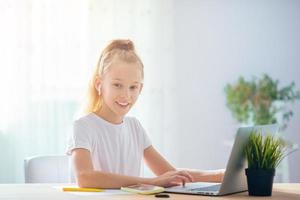 Serious schoolgirl sitting at table with laptop and textbook and doing homework. photo