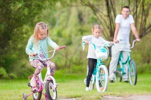 niñas adorables y papá feliz andando en bicicleta en un día cálido de verano. paseo familiar joven activo en bicicleta foto