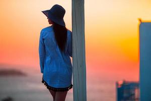 Young woman is looking at the sunset over a sea in famous village Mykonos with the old buildings in the background. Soft summer backlight. photo