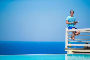 Caucasian guy relaxing near pool with amazing view and reading book in Greece. Beautiful famous background photo