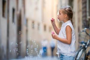 Adorable little girl outdoors blowing soap bubbles in european city. Portrait of caucasian kid enjoy summer vacation in Italy photo