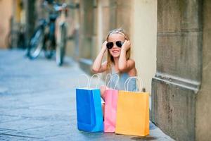 retrato de una adorable niña caminando con bolsas de compras al aire libre en una ciudad europea. niño pequeño de moda en la ciudad italiana con sus compras foto